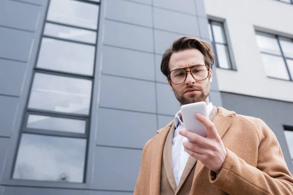 Low angle view of bearded businessman in glasses and beige coat using cellphone near building outside — Stock Photo