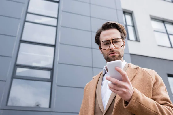 Vue à angle bas de l'homme d'affaires barbu en lunettes et manteau beige à l'aide d'un téléphone portable près du bâtiment à l'extérieur — Photo de stock
