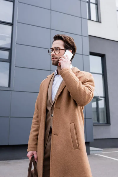 Vista de ángulo bajo del hombre de negocios barbudo en gafas y abrigo beige hablando en el teléfono celular cerca del edificio - foto de stock