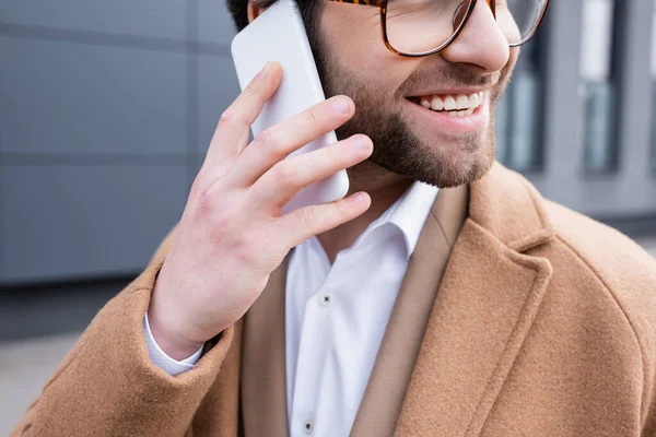 Vista recortada de hombre de negocios feliz en gafas y abrigo beige hablando en el teléfono celular cerca del edificio exterior - foto de stock