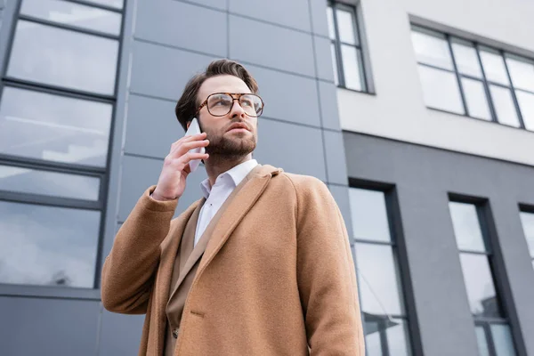 Low angle view of man in glasses and beige coat talking on cellphone near building outside — Stock Photo