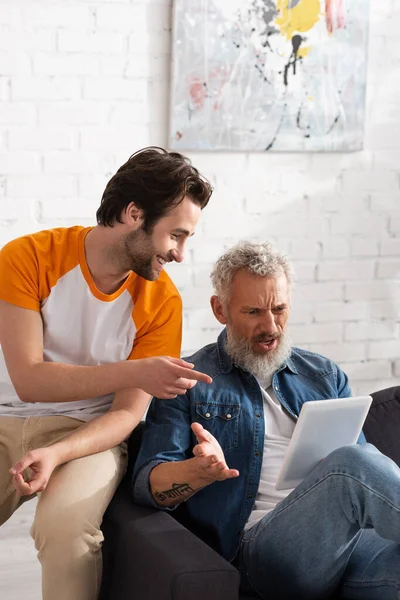 Smiling man pointing with finger at digital tablet near angry father — Stock Photo