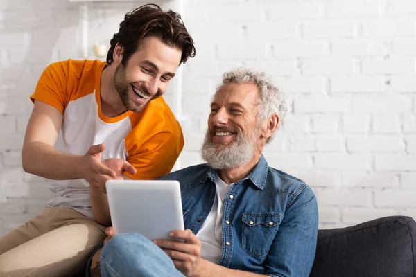 Uomo sorridente che punta al tablet digitale vicino al padre maturo a casa — Foto stock