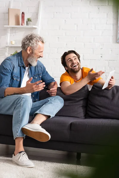 Smiling man pointing at digital tablet near middle aged father on couch — Stock Photo