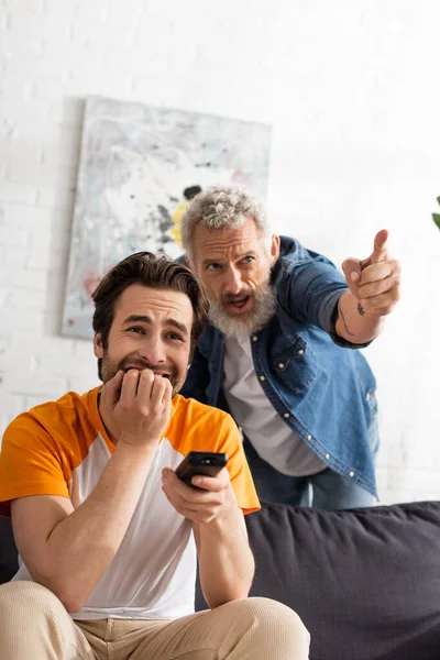 Hombre preocupado sosteniendo mando a distancia cerca de padre señalando con el dedo - foto de stock