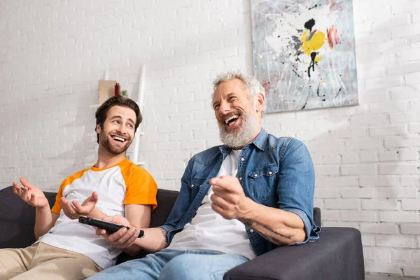 Homme joyeux avec télécommande pointant avec la main près du fils sur le canapé — Photo de stock