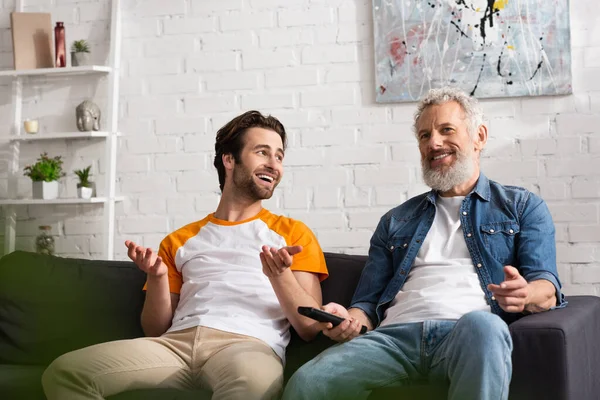 Smiling man looking at mature father with remote controller on couch — Stock Photo