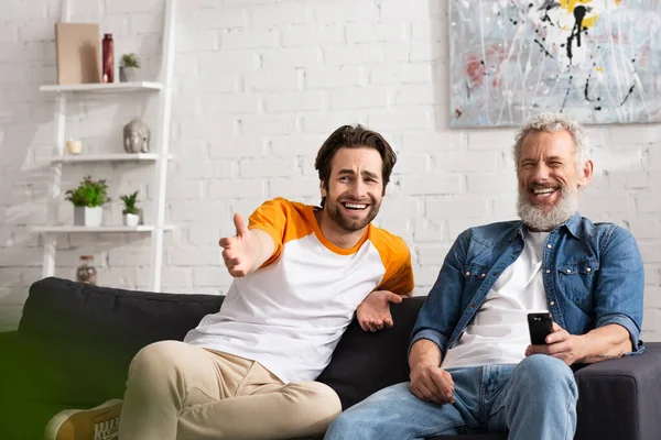 Homme souriant pointant avec la main près du père avec télécommande sur le canapé — Photo de stock