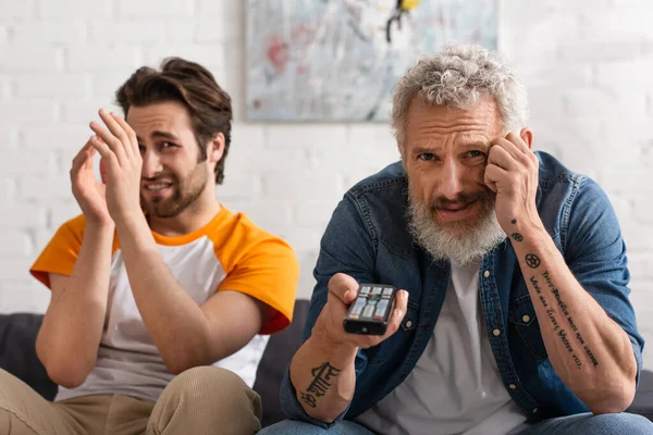 Hombre preocupado sosteniendo mando a distancia cerca borrosa hijo en el sofá - foto de stock