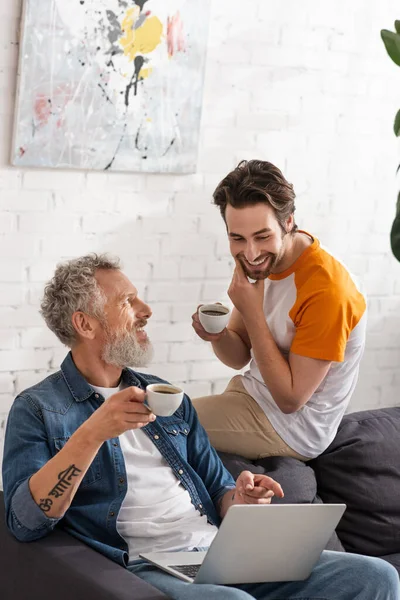 Homme positif et fils tenant des tasses à café près d'un ordinateur portable sur le canapé — Stock Photo