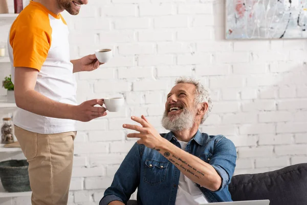 Lächelnder älterer Mann sitzt auf Couch neben Sohn mit Tassen Kaffee — Stockfoto