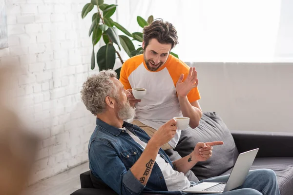 Mature man pointing at laptop near son with coffee on couch — Stock Photo