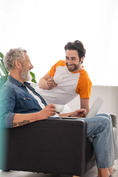 Sonriente padre e hijo sosteniendo tazas de café cerca de la computadora portátil - foto de stock