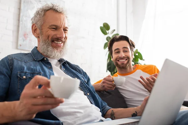 Uomo sorridente che tiene il caffè vicino al figlio e al computer portatile offuscato — Foto stock