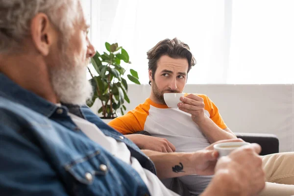 Hombre con café mirando a padre en primer plano borroso en casa - foto de stock