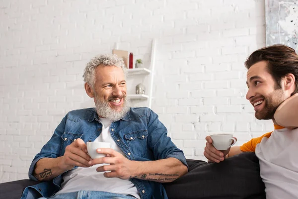 Молодой человек разговаривает с улыбающимся отцом, держа чашку кофе в гостиной — стоковое фото