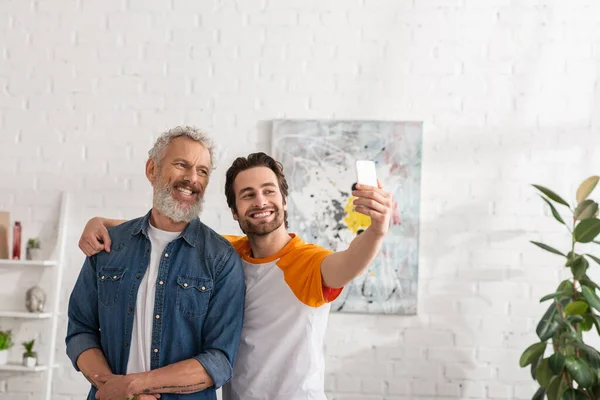 Hijo abrazando padre y tomando selfie con smartphone en la sala de estar - foto de stock