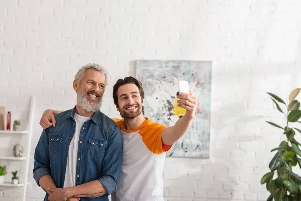 Fils étreignant père et prendre selfie avec smartphone dans le salon — Photo de stock