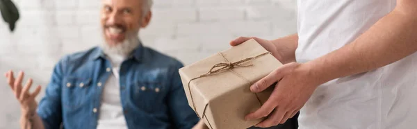 Чоловік тримає подарункову коробку біля розмитого батька вдома, банер — стокове фото