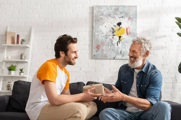 Smiling son holding gift box near father on couch — Stock Photo
