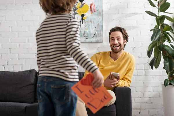 Lächelnder Mann mit Smartphone schaut Sohn mit Geschenkkarte an — Stockfoto