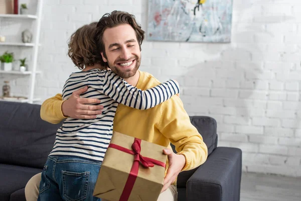 Sonriente hombre abrazando hijo y sosteniendo presente en casa - foto de stock