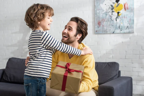 Enfant joyeux embrassant père avec présent sur le premier plan flou — Photo de stock