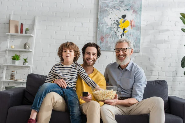 Smiling grandfather holding bowl with popcorn near son with remote controller and grandson — Stock Photo