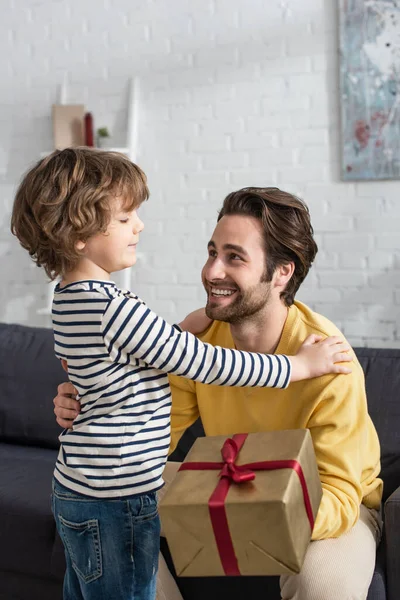 Sonriente padre con caja de regalo sentado cerca de su hijo en casa - foto de stock