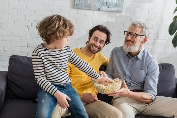 Kind nimmt Popcorn neben lächelndem Vater und Großvater zu Hause — Stockfoto