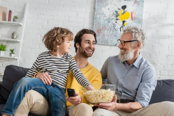Sorrindo pai com controlador remoto e avô com pipoca perto da criança no sofá — Fotografia de Stock