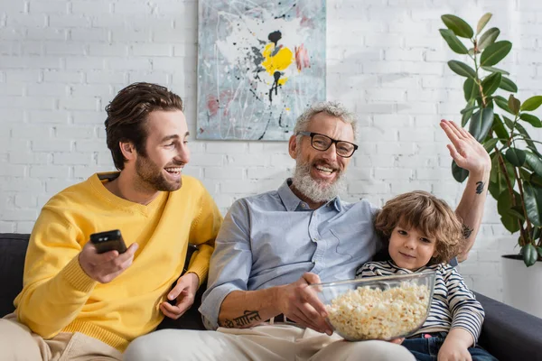 Uomo sorridente con popcorn mano ondulante vicino al figlio con telecomando e nipote sul divano — Foto stock