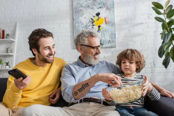 Men and child sitting with popcorn and remote controller on couch — Stock Photo