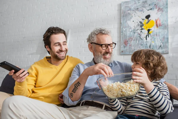Sonriente hombre con mando a distancia sentado cerca de padre e hijo con palomitas de maíz en el sofá - foto de stock