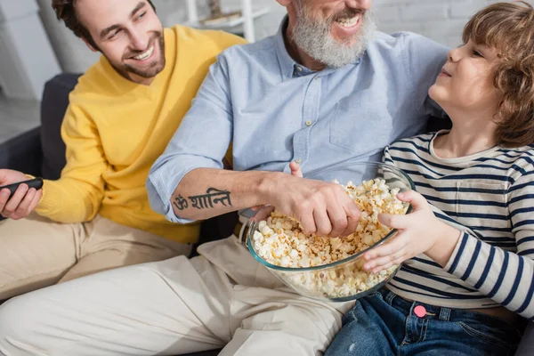 Lächelnder Junge hält Popcorn in der Nähe von Großvater und Vater mit Fernbedienung — Stockfoto