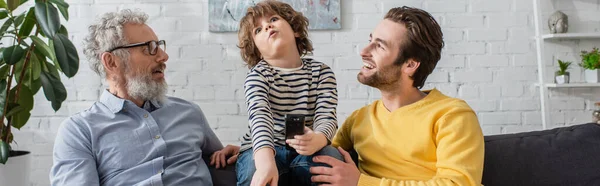 Sonriente padre y abuelo mirando al niño con mando a distancia, pancarta - foto de stock