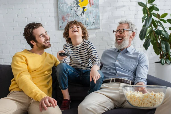 Hombres felices con palomitas de maíz sentados cerca de niño con mando a distancia - foto de stock