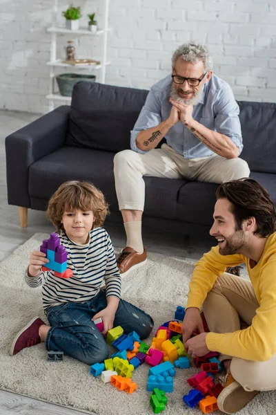 Niño sosteniendo bloques de construcción cerca de padre y abuelo en casa - foto de stock