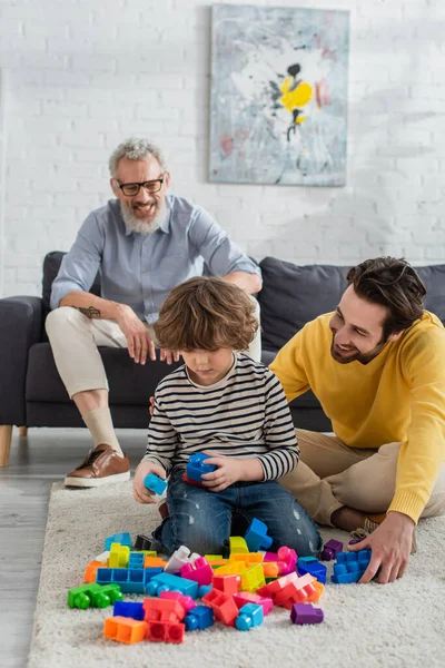 Cheerful man sitting near son playing building blocks and blurred father — Stock Photo