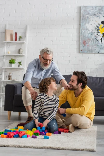 Reifer Mann sitzt neben lächelndem Enkel und Sohn und spielt Bauklötze — Stockfoto