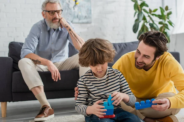 Smiling man holding building block near son and blurred father at home — Stock Photo