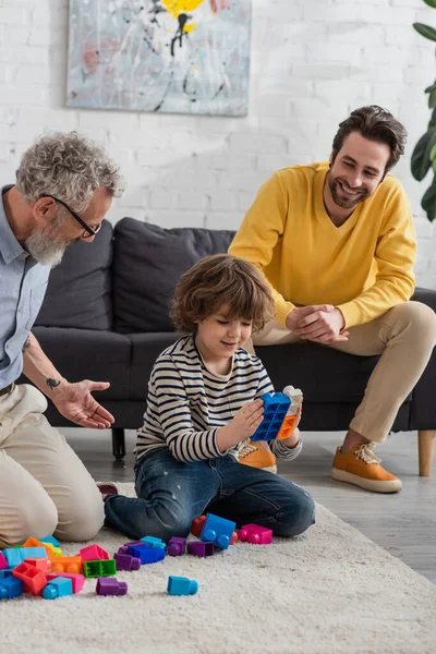 Smiling kid playing building blocks near father and grandfather at home — Stock Photo