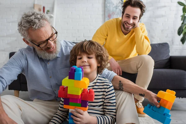 Abuelo con bloques de construcción sentado cerca de niño e hijo sonriente sobre fondo borroso - foto de stock