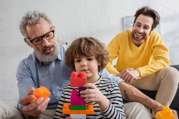 Niño jugando bloques de construcción cerca del abuelo y el padre borroso - foto de stock