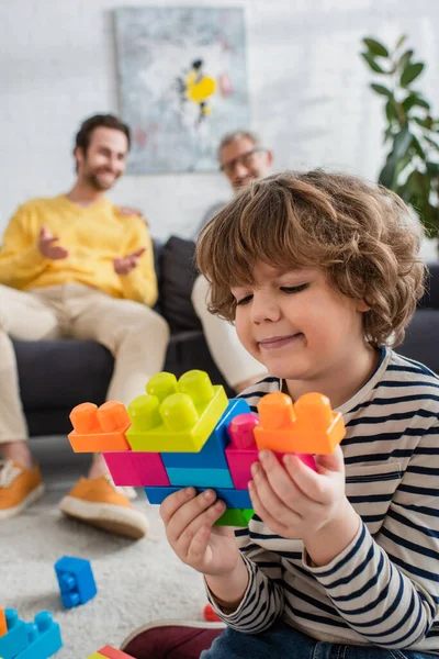 Smiling boy playing building blocks near father and grandfather on blurred background — Stock Photo