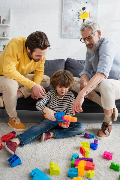 Uomo maturo e figlio seduto vicino nipote con blocchi di costruzione al piano — Foto stock