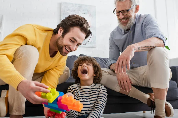 Aufgeregter Junge sitzt neben lächelnden Eltern mit verschwommenen Bauklötzen zu Hause — Stockfoto