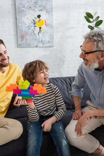 Sorridente ragazzo tenendo blocchi di costruzione vicino nonno e padre sul divano — Foto stock