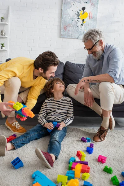 Positive parents looking at kid near building blocks on carpet — Stock Photo