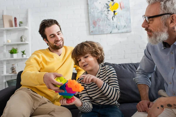 Boy pointing at building blocks near smiling father and grandfather on couch — Stock Photo